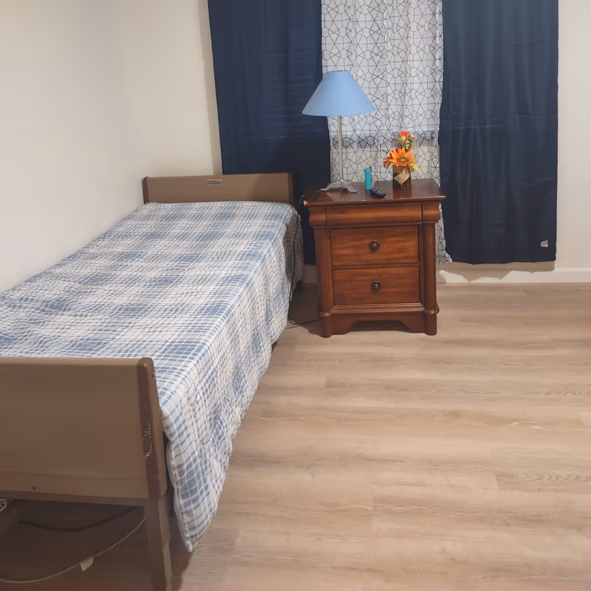 Simple bedroom with a single bed covered in a blue plaid bedspread next to a wooden nightstand holding a lamp and small decor in front of dark curtains.