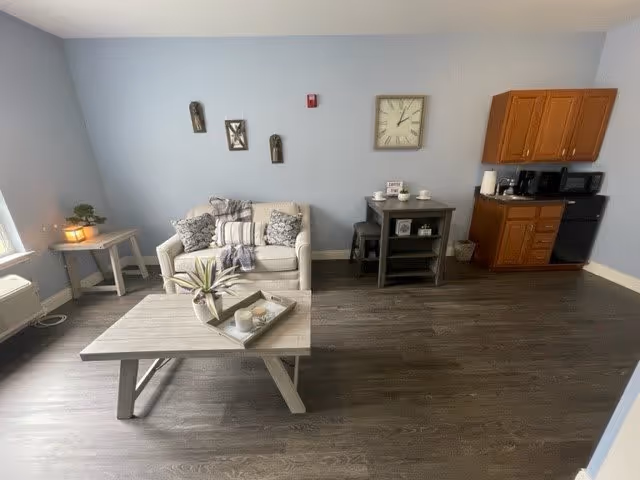 A cozy living area with a light gray loveseat adorned with patterned pillows and a throw blanket. In front of the loveseat is a wooden coffee table with a decorative tray holding candles and a small plant. To the left, there is a small side table with a lit lantern and a potted plant. On the right side of the room, there is a small kitchenette with wooden cabinets, a microwave, a coffee maker, and a mini refrigerator. Above a small black table with stools on the right wall, there is a large clock and some decorative items. The walls are painted light blue and the floor is dark wood.