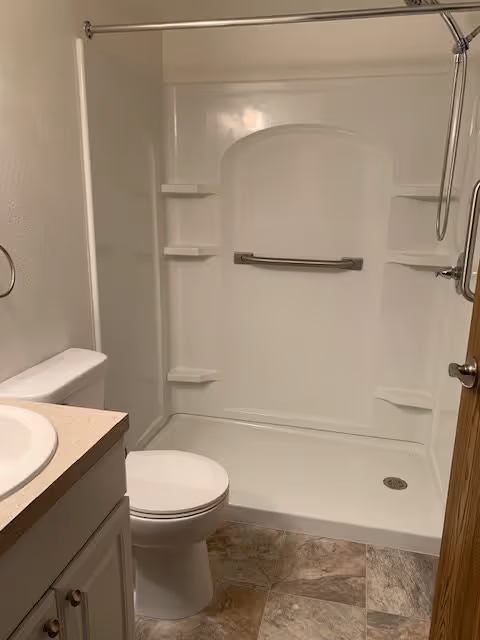 A small bathroom featuring a white toilet, a white sink with a beige countertop, and a white shower with built-in shelves and a grab bar. The floor has a tile pattern in neutral tones, and a shower curtain rod is installed above the shower.
