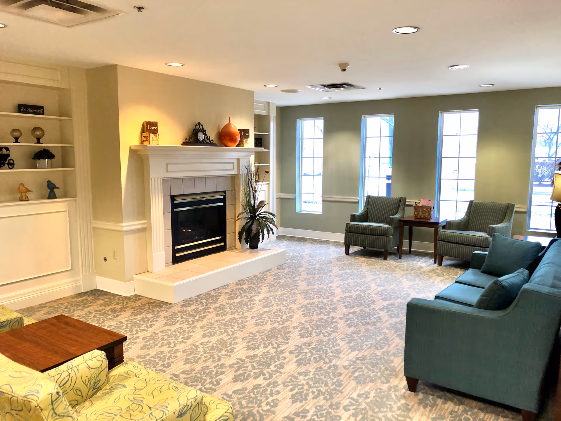 A cozy living room area in an assisted living facility featuring a fireplace with decorative items on the mantel, built-in shelves with small decor pieces, patterned carpet, multiple armchairs, a sofa, and large windows letting in natural light.