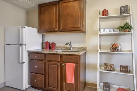 A small kitchen area with a white refrigerator on the left, wooden cabinets above and below a granite countertop with a sink. A red towel hangs from the cabinet handle. To the right, a white ladder-style shelf holds decorative items including a red container, a sign that says 'Bless this home', a glass bowl with decorative balls, and small wooden blocks with words like 'Live' and 'Love'.