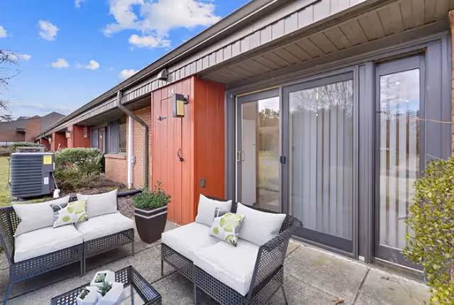 Outdoor patio area at Resorts of Augsburg featuring two cushioned wicker loveseats with decorative pillows, a small glass-top table with plants, potted plants, and sliding glass doors leading inside. The building exterior is brick with red and gray paneling under a partly cloudy sky.