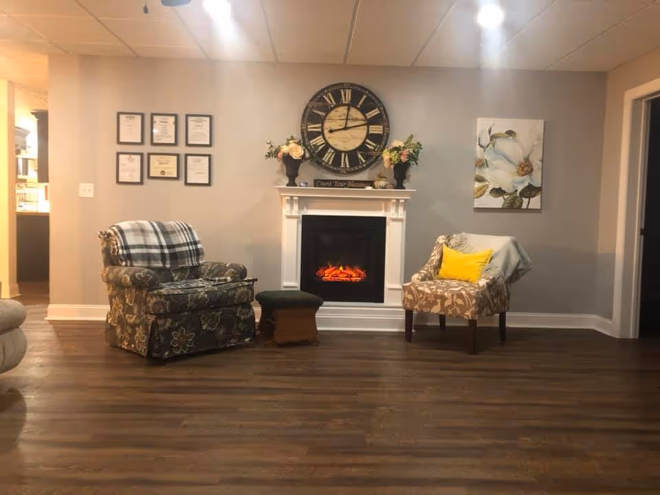 A cozy sitting area with two armchairs flanking a lit electric fireplace topped by a large wall clock and floral decorations.