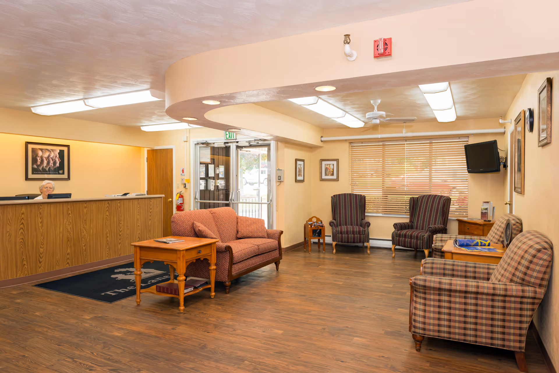 A senior living facility lobby with a wooden reception desk where a staff member is seated. The room features a wooden floor, a red patterned sofa, a wooden coffee table, several armchairs with striped and plaid upholstery, a wall-mounted TV, and framed artwork on the walls. Large windows with blinds allow natural light to enter the space.