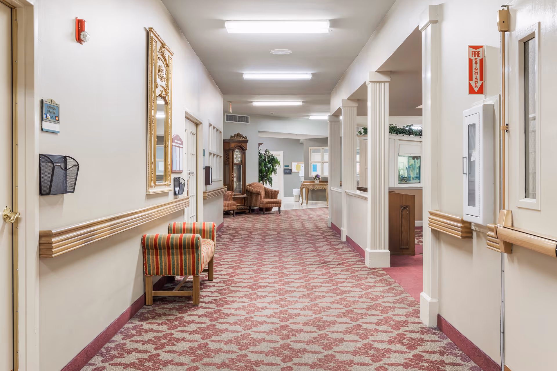 A well-lit hallway in a senior living facility with patterned carpet, beige walls, and wooden handrails. There are chairs along the walls, a large ornate mirror, a grandfather clock, and some plants visible in the distance. The hallway leads to a common area with columns and additional seating.