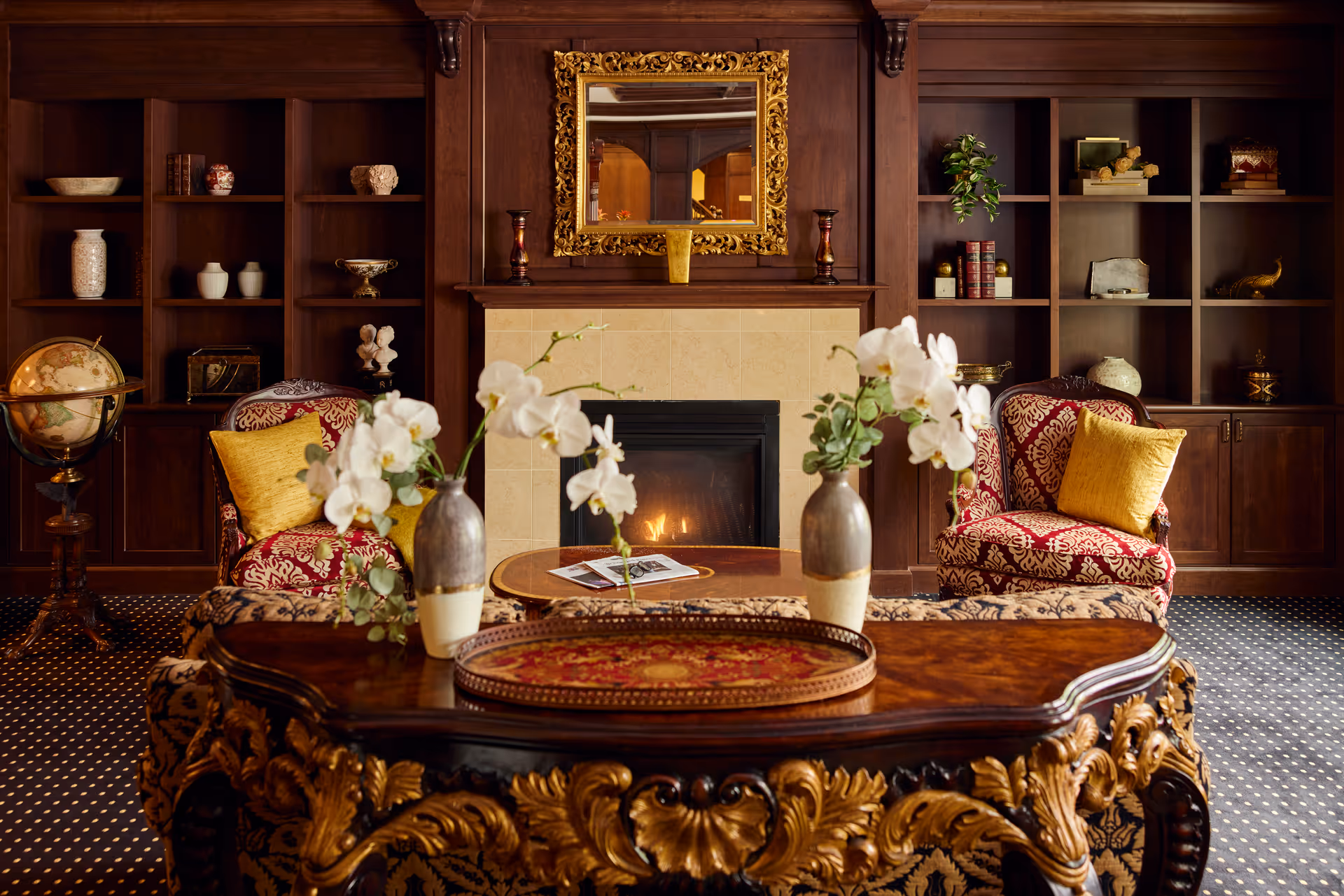 A cozy living room with a lit fireplace surrounded by dark wood paneling and built-in shelves. Two patterned armchairs with yellow cushions flank the fireplace. A decorative wooden table with intricate carvings holds two vases with white orchids in the foreground. A gold-framed mirror hangs above the fireplace.