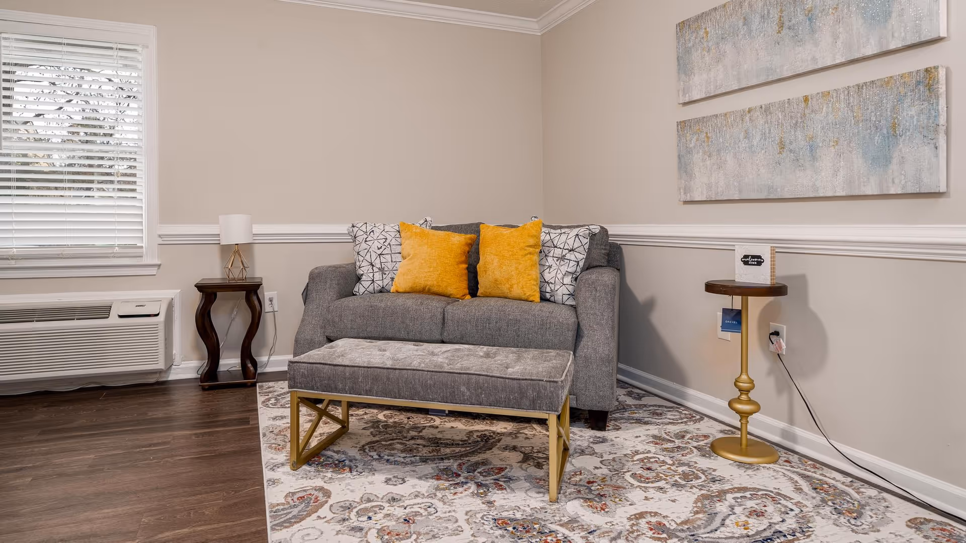A cozy living room area featuring a gray loveseat with two yellow and two patterned throw pillows, a matching gray ottoman with gold legs, a patterned area rug, a small wooden side table with a lamp, and a round gold side table with a small sign. The walls are beige with white trim, and there are two abstract paintings hanging above the loveseat. A window with white blinds and an air conditioning unit are visible on the left side.