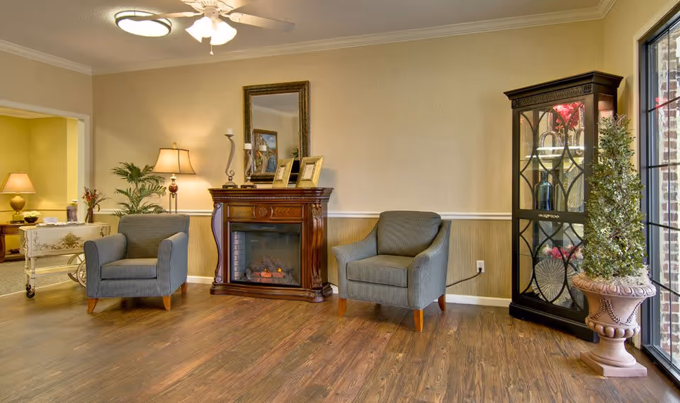 A cozy living room area with two gray armchairs facing a wooden electric fireplace. Above the fireplace is a mirror and decorative items including picture frames and candles. To the right is a glass display cabinet with decorative items inside and a potted plant next to it. The room has wooden flooring, beige walls, and a ceiling fan with lights. A large window on the right lets in natural light.
