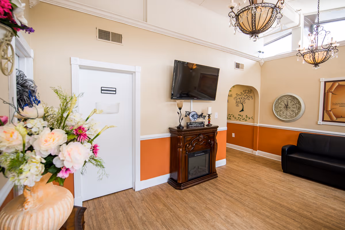 A cozy interior room with beige and orange walls, a wooden floor, a black leather couch, a wall-mounted flat screen TV above a decorative wooden electric fireplace, a large round clock on the wall, floral decorations in a vase, and two ornate hanging light fixtures.