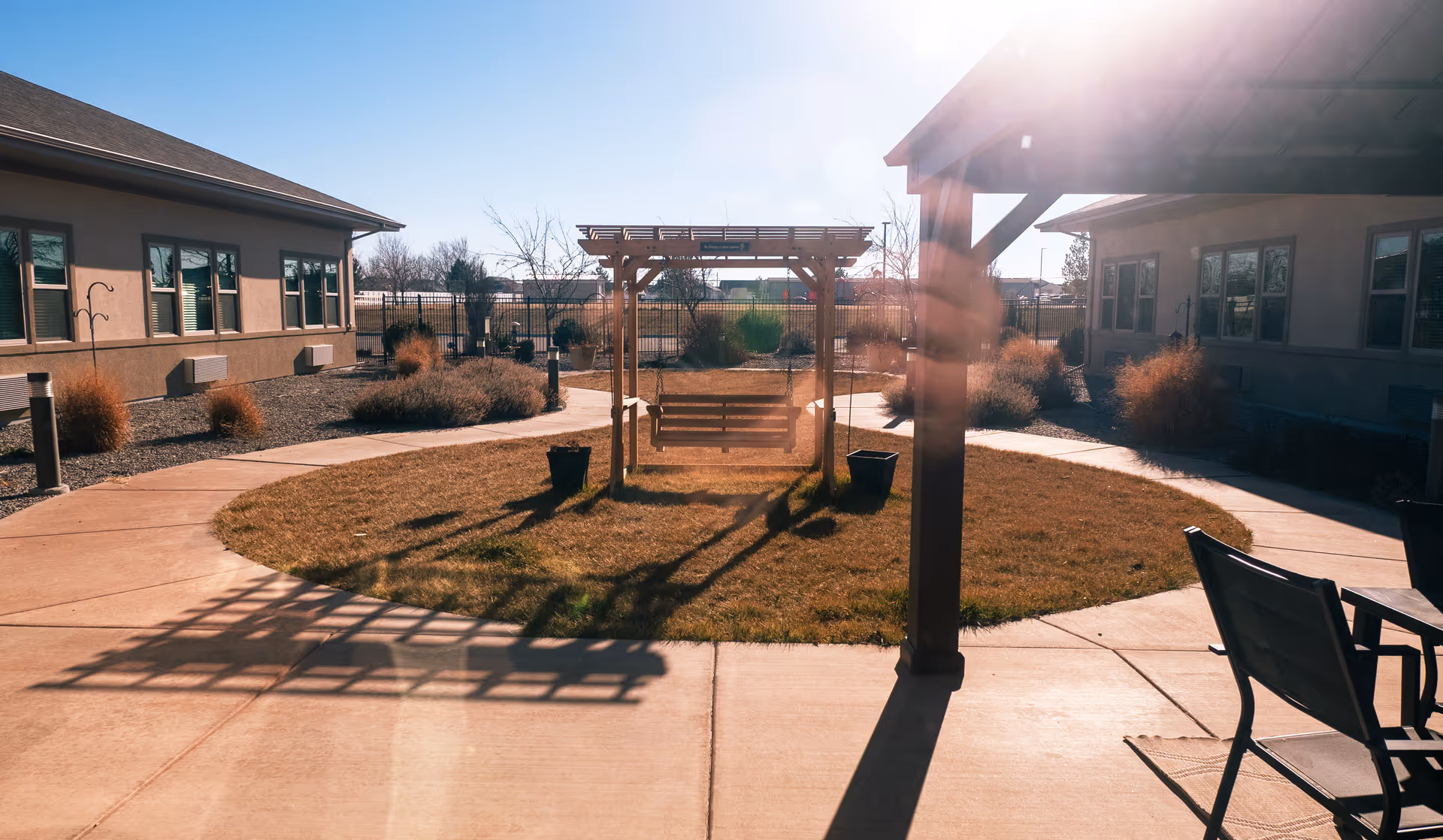 Outdoor courtyard area at a senior living facility with a wooden swing under a pergola, surrounded by a circular patch of grass and concrete walkways. There are two buildings on either side with windows, some shrubs, and outdoor chairs and tables visible on the right side.