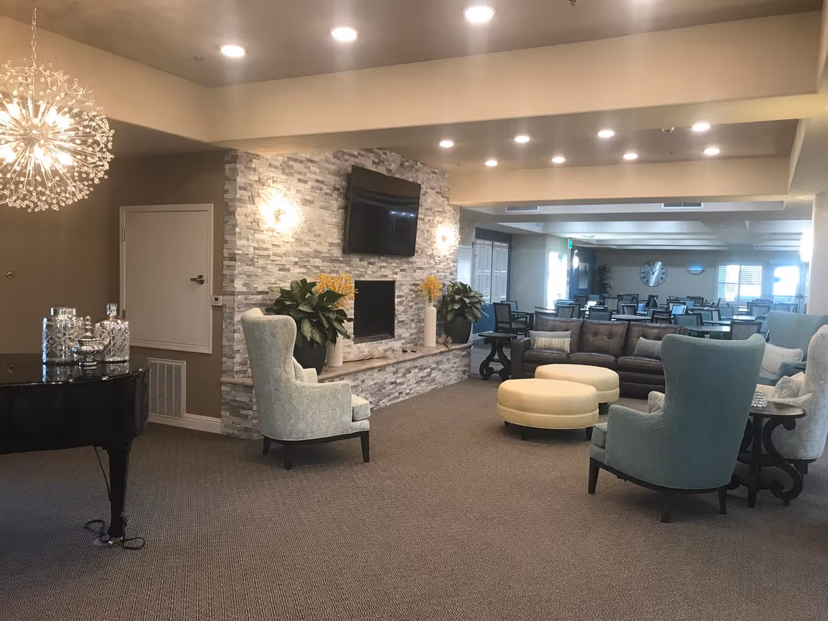 Open, well-lit senior living common area with armchairs, ottomans, a fireplace and mounted TV, and dining tables visible in the background.