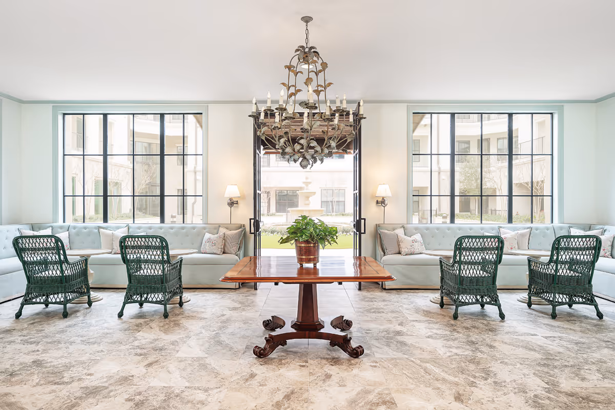 Bright and spacious living room with large windows overlooking an outdoor courtyard. The room features two long light blue tufted sofas along the walls, four green wicker chairs arranged in pairs facing each other, a wooden table with a potted plant in the center, and a decorative chandelier hanging from the ceiling.