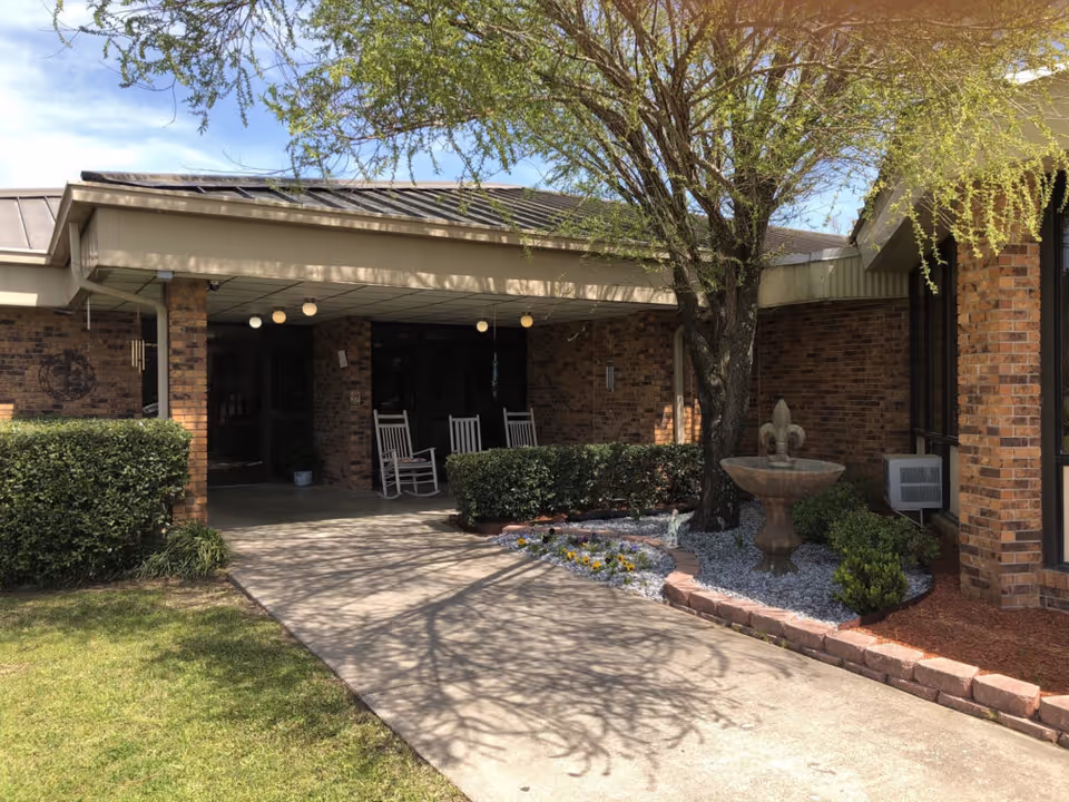 Entrance area of a brick building with a covered walkway, three white rocking chairs, a tree, a small garden with flowers and a decorative fountain, and trimmed bushes under a partly cloudy sky.