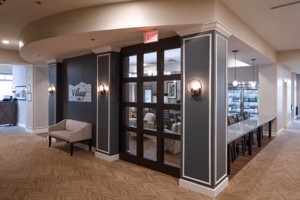 Lobby corridor showing the entrance to the Village Café with glass-paneled doors, a bench, wall sconces, and a long counter with stools and refrigerated displays.