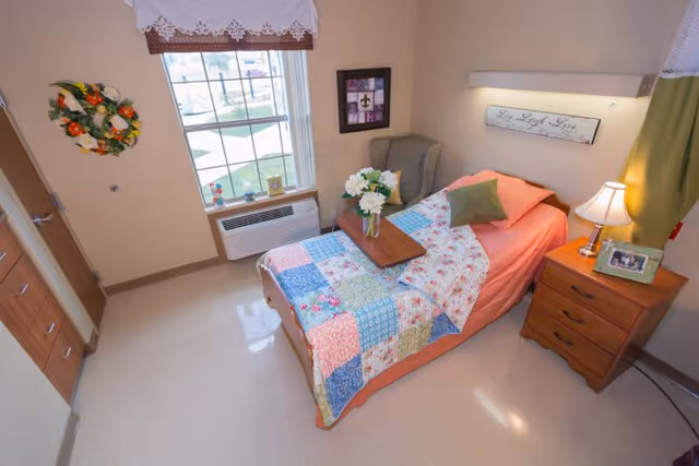 A small, cozy bedroom in a senior living facility with a single bed covered in a colorful patchwork quilt. There is a wooden nightstand with a lamp and a framed photo, a cushioned armchair in the corner, and a window with a white valance letting in natural light. A small table with a vase of white flowers is placed on the bed.