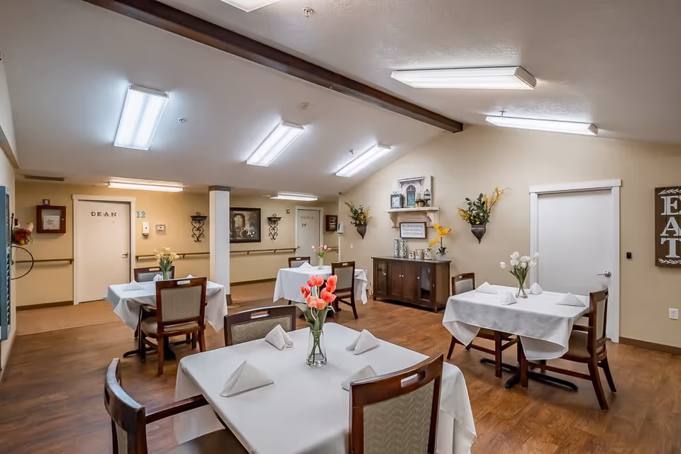 Dining room with small tables set with white tablecloths and flower centerpieces in a memory care facility.