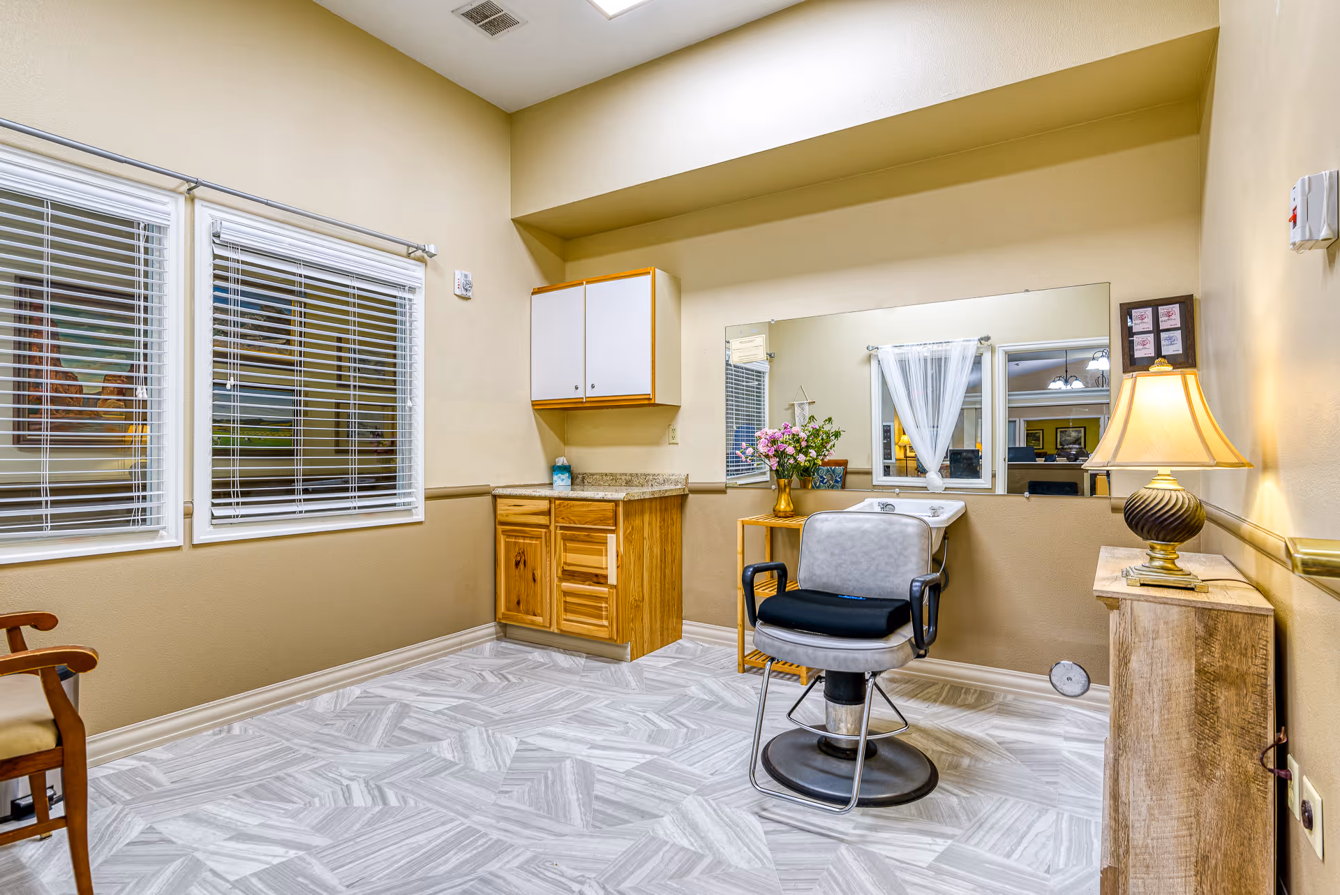 A well-lit room with beige walls and patterned gray floor tiles, featuring a salon chair in front of a large mirror mounted on the wall. To the left, there are two windows with white blinds and a wooden cabinet with a countertop. On the right side, a wooden side table holds a lamp with a beige shade, and a small vase with flowers is placed near the mirror.