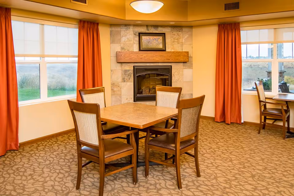 A cozy interior room with a square wooden table surrounded by four chairs with cushioned seats and backs. The room features a fireplace with a stone surround and a wooden mantel, above which hangs a framed picture. Large windows with orange curtains allow natural light to enter, and the carpet has a patterned design.