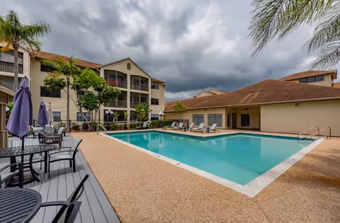 Outdoor swimming pool area at Waterside Landing with lounge chairs and tables with umbrellas around the pool. The pool is surrounded by a beige textured deck, and the building with balconies and palm trees is visible in the background under a cloudy sky.