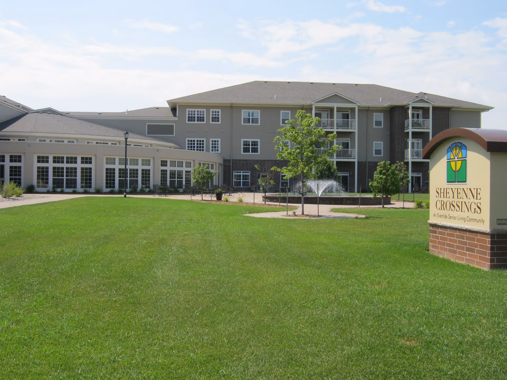 Exterior view of Sheyenne Crossings senior living community building with a well-maintained green lawn, small trees, a water fountain, and a sign displaying the community name.