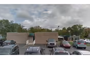Parking lot in front of a single-story beige building with a flat roof under a cloudy sky. Several cars are parked in the lot, and there are trees in the background.