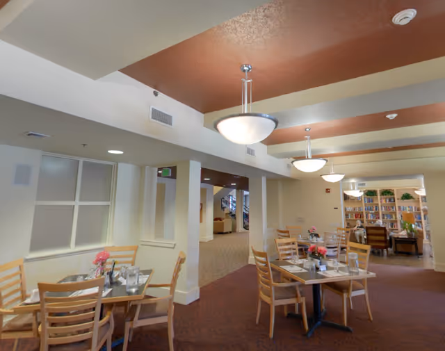 Interior view of a dining area in an assisted living facility with several wooden tables and chairs. Each table is set with glasses, napkins, and small flower arrangements. The ceiling has modern light fixtures, and in the background, there is a cozy seating area with bookshelves filled with books.