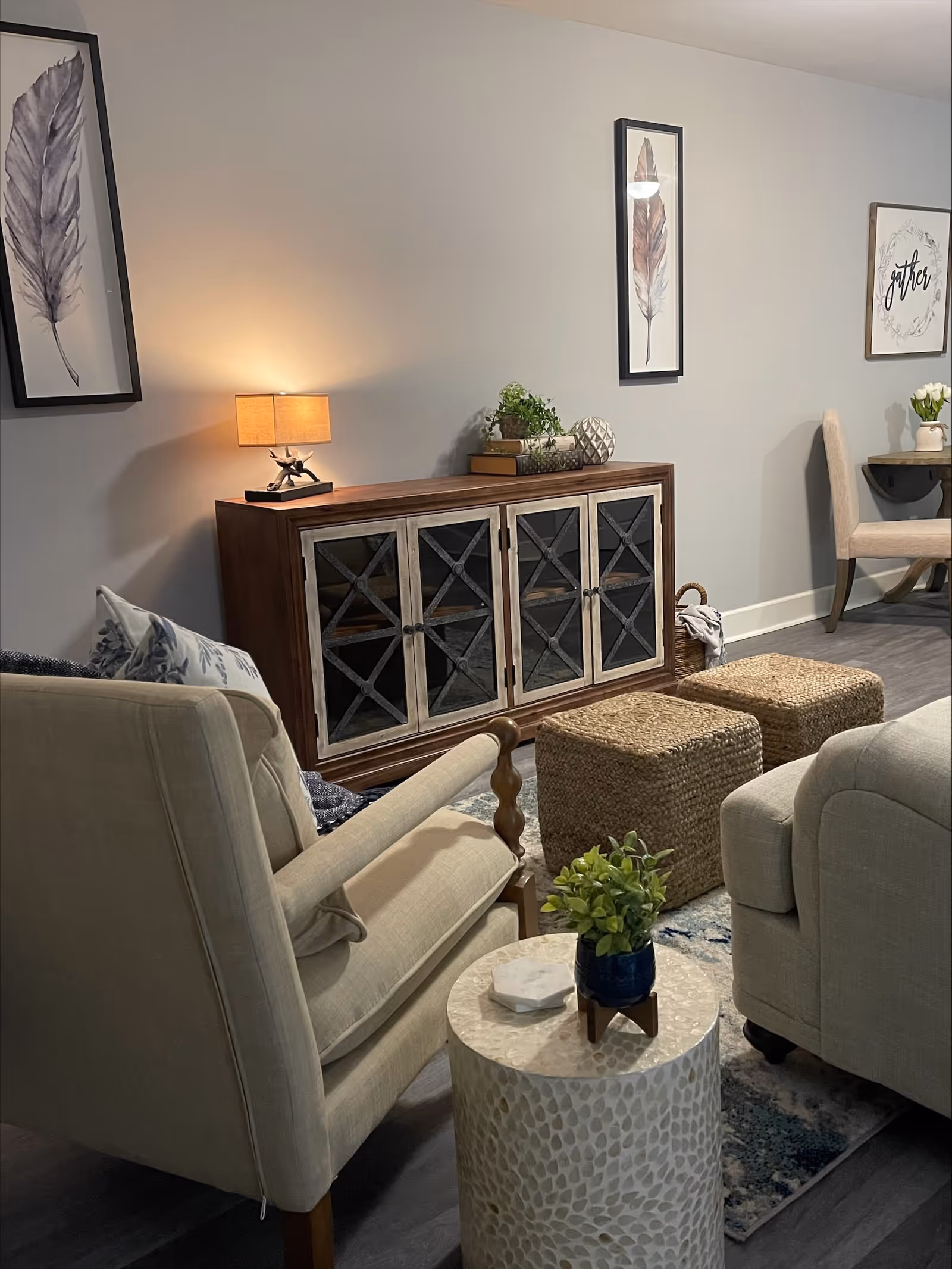 A cozy living room area with beige upholstered armchairs, a small round side table with a potted plant, two woven ottomans, and a wooden cabinet with glass-paneled doors. The wall behind the cabinet features framed artwork of feathers and a sign that says 'gather'. A small table lamp is placed on the cabinet, casting a warm light.