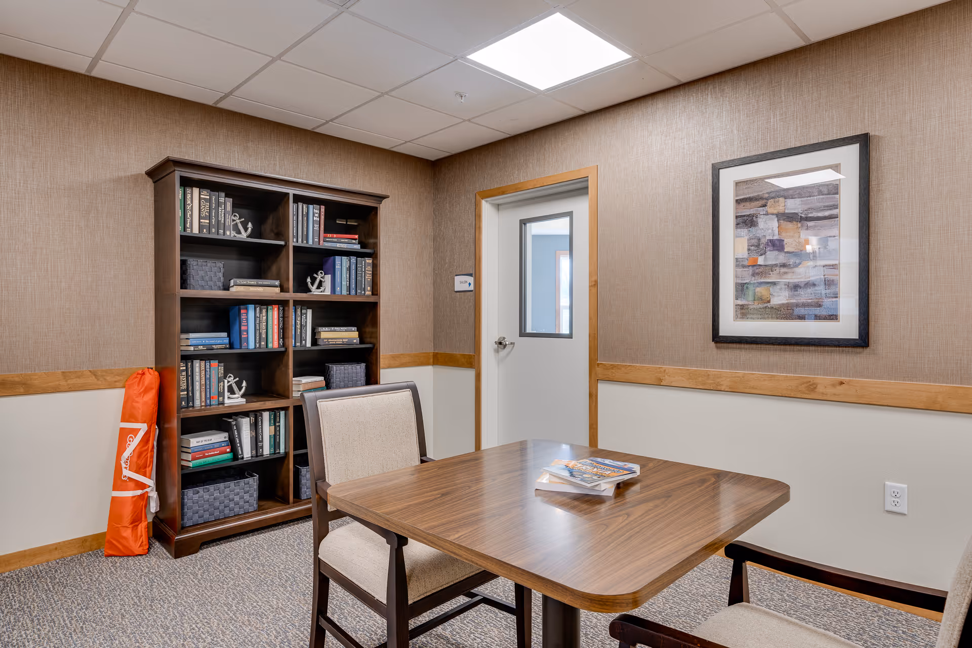 A small room with a wooden table and two upholstered chairs. There is a bookshelf filled with books and decorative items against one wall, and a framed abstract painting on another wall. A door with a window is visible in the background. The room has beige walls with wooden trim and a carpeted floor.