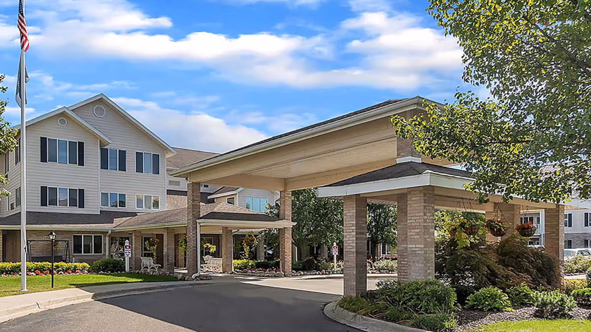 Exterior view of Holiday Genesee Gardens facility showing a covered entrance with brick pillars, landscaped greenery, and a three-story building with multiple windows under a partly cloudy sky.