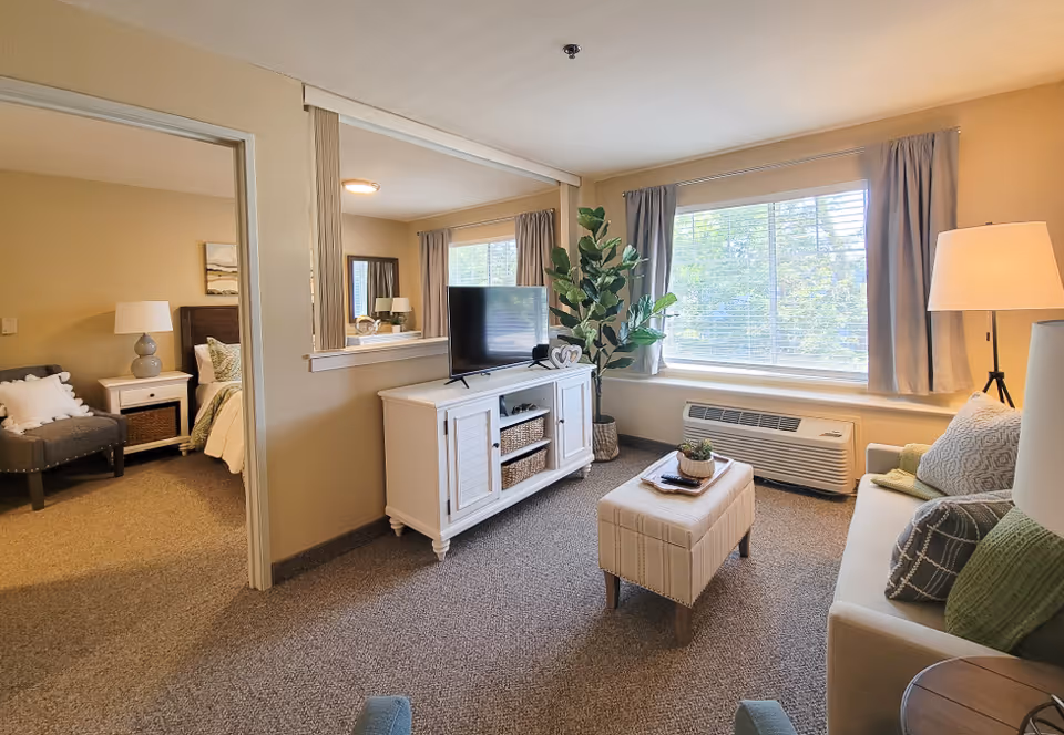 Bright living room with a sofa, ottoman, TV on a white console, large window and a view into an adjacent bedroom.