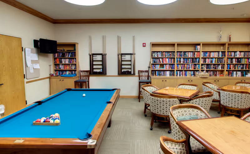 A recreational room with a pool table set up for a game, several wooden tables with cushioned chairs, bookshelves filled with books, and pool cues mounted on the wall. The room has beige walls, a carpeted floor, and a wooden door on the left side.