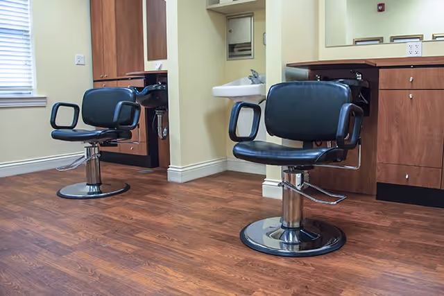 Interior view of a salon area in Richmond Heights Place featuring two black salon chairs with armrests on a wooden floor, a small sink mounted on the wall, and wooden cabinetry with drawers and a mirror.