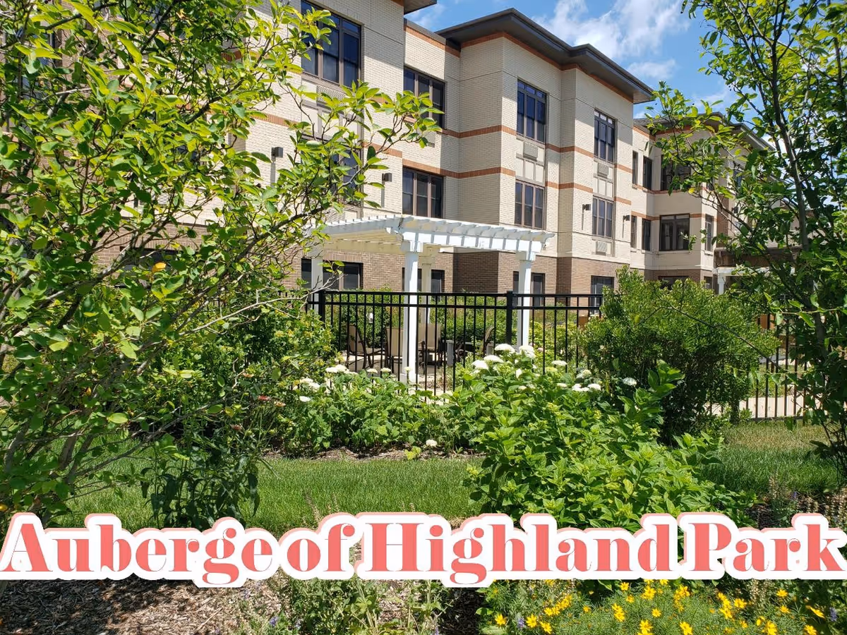 Outdoor garden area with green shrubs, flowering plants, and a white pergola with seating, adjacent to a multi-story brick building under a blue sky with some clouds.