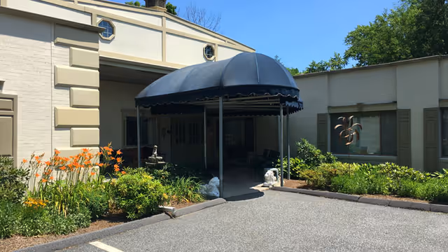 Entrance to a building with a black canopy over the doorway, surrounded by flower beds with orange and yellow flowers and green shrubs. The building is light-colored with decorative trim and has a paved driveway in front.