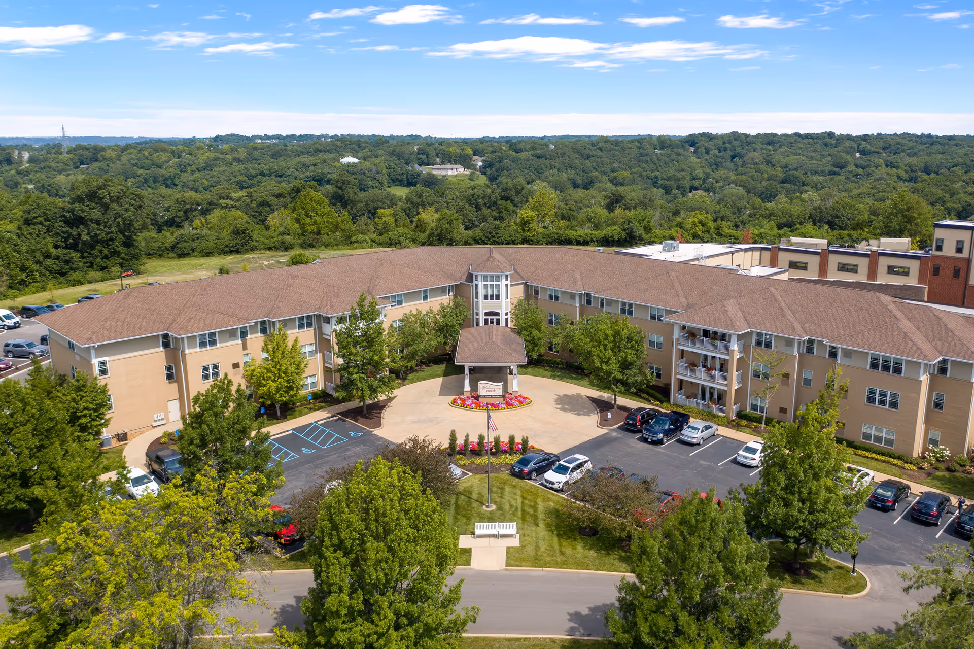 Aerial view of Friendship Village Sunset Hills, a large senior living facility building with a brown roof and beige exterior walls. The building is surrounded by green trees and a parking lot with several cars. There is a circular driveway with a covered entrance and a landscaped area with flowers and an American flag in front of the building. The background shows a vast expanse of green forested area under a partly cloudy blue sky.