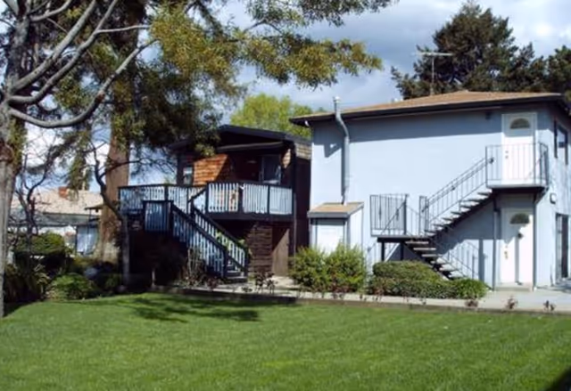 Exterior view of a two-story building with outdoor staircases leading to the upper floors, surrounded by green grass, bushes, and trees under a partly cloudy sky.