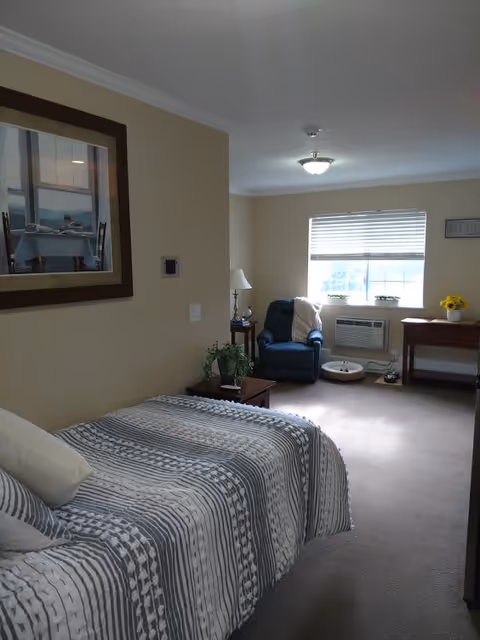 A cozy bedroom in an assisted living facility featuring a bed with striped bedding, a framed picture on the wall, a small wooden side table with a plant and lamp, a blue armchair near a window with blinds, an air conditioning unit below the window, and a wooden desk with a vase of yellow flowers.