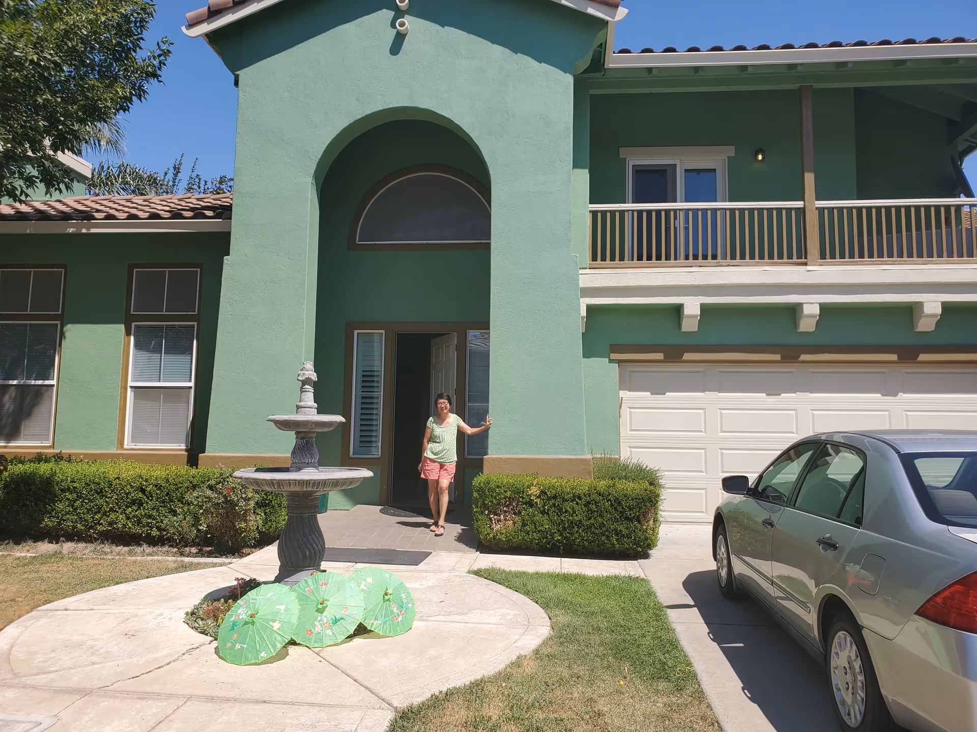 A woman standing at the entrance of a green two-story house with a tiled roof. There is a two-tiered fountain in front of the house and a silver car parked in the driveway. The house has several windows and a balcony above the garage.
