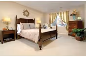 Spacious bedroom with a wooden bed, nightstands with lamps, a dresser, window with curtains, and potted plants.