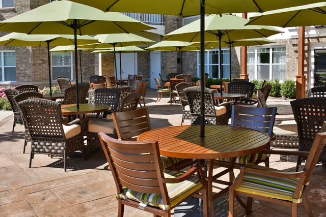 Outdoor patio area with multiple round wooden tables and cushioned chairs under large green umbrellas, surrounded by a building with windows and some greenery.