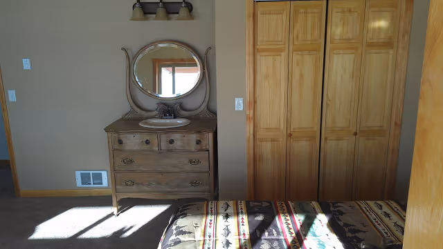 Sunny bedroom with a wooden dresser topped by a round mirror, built-in wooden closet doors, and a patterned bedspread.