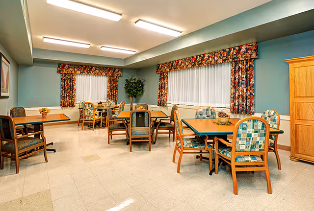 A dining room with multiple square wooden tables and cushioned chairs with patterned upholstery. The walls are painted light blue with floral curtains on the windows. There is a wooden cabinet on the right side and a potted plant near the windows.