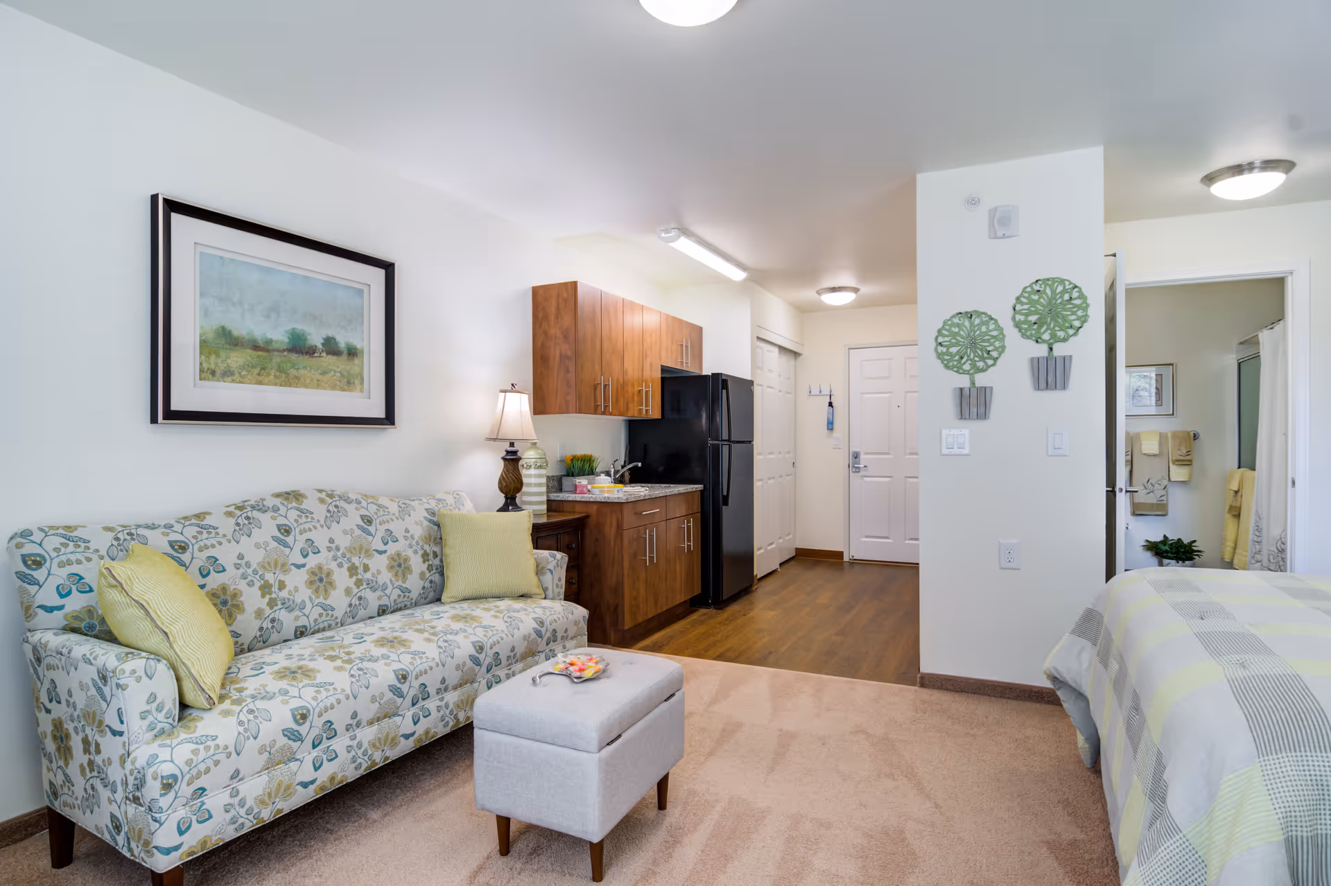 A cozy studio apartment interior featuring a floral patterned sofa with yellow cushions, a small gray ottoman, a kitchenette with wooden cabinets and a black refrigerator, a bed with a checkered bedspread, and a bathroom visible through an open door. The walls are decorated with framed artwork and green plant wall hangings.