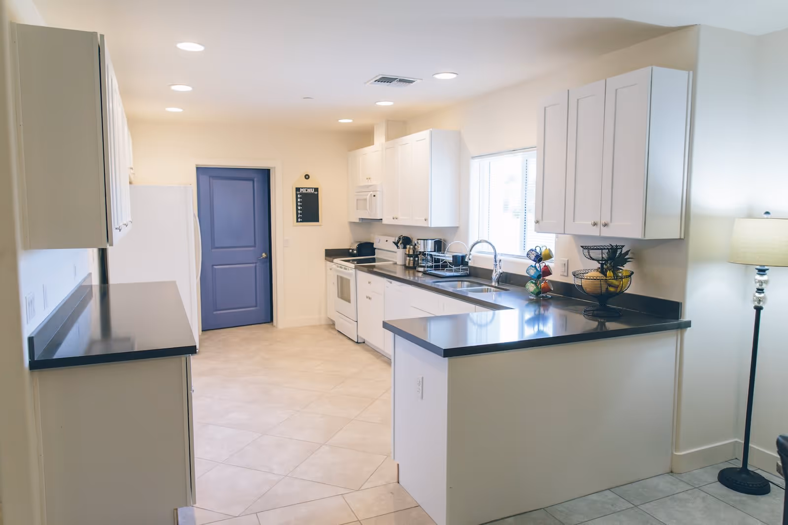 A modern kitchen with white cabinets, black countertops, a white stove, microwave, dishwasher, and refrigerator. There is a blue door at the end of the kitchen and a window above the sink letting in natural light. A floor lamp is visible on the right side.