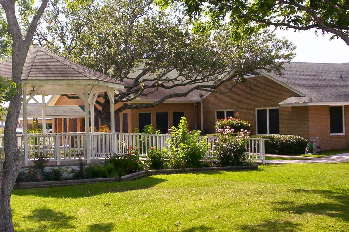 Outdoor view of a senior living facility with a white gazebo, green lawn, trees, and a brick building in the background.