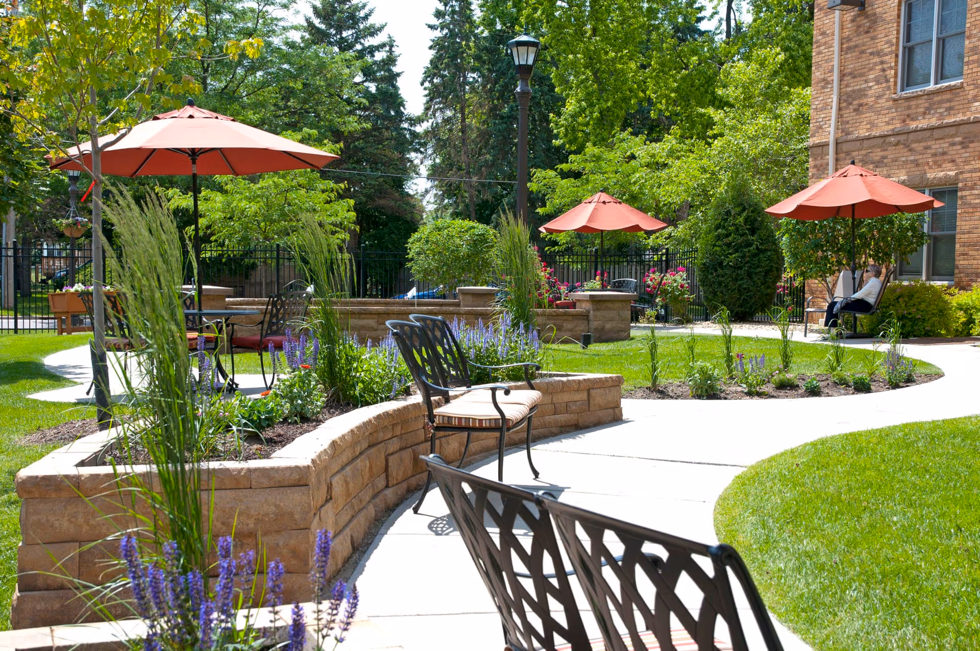 A sunny outdoor garden area at The Alton with curved stone planters filled with flowers and tall grasses. There are several black metal chairs with cushions and tables shaded by red umbrellas. A paved walkway winds through the green lawn and landscaping. A person is sitting on a bench near the brick building in the background.