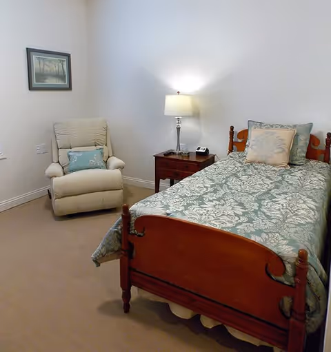 A cozy bedroom in an assisted living community featuring a wooden bed with a floral patterned bedspread and multiple pillows. Next to the bed is a wooden nightstand with a lamp and a phone. There is a beige recliner chair with a decorative pillow in the corner, and a framed picture hanging on the wall above it.