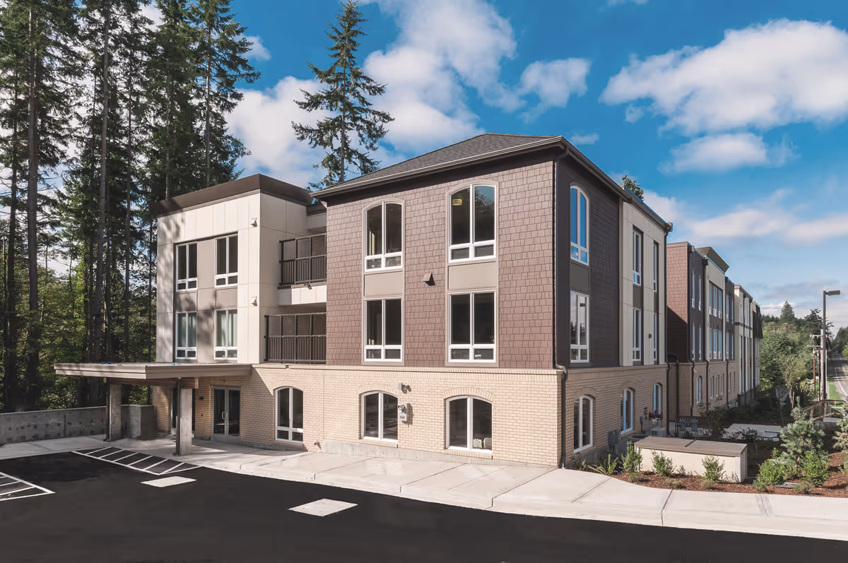 Exterior front view of a three-story senior living building with brick and siding, an entrance canopy, parking area, and trees in the background.