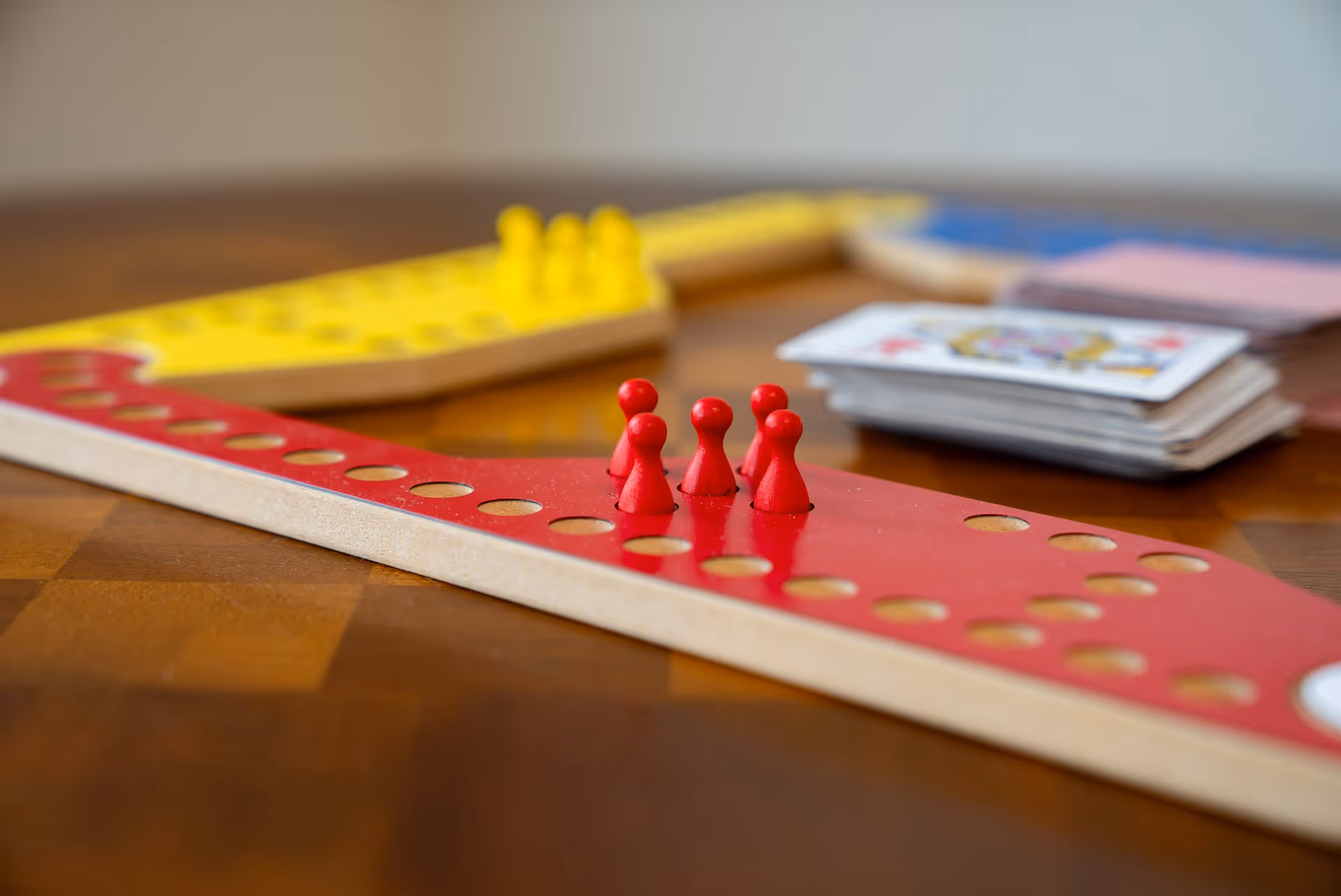 Close-up of a wooden board game with red and yellow playing pieces on a wooden table, with a stack of playing cards in the background.