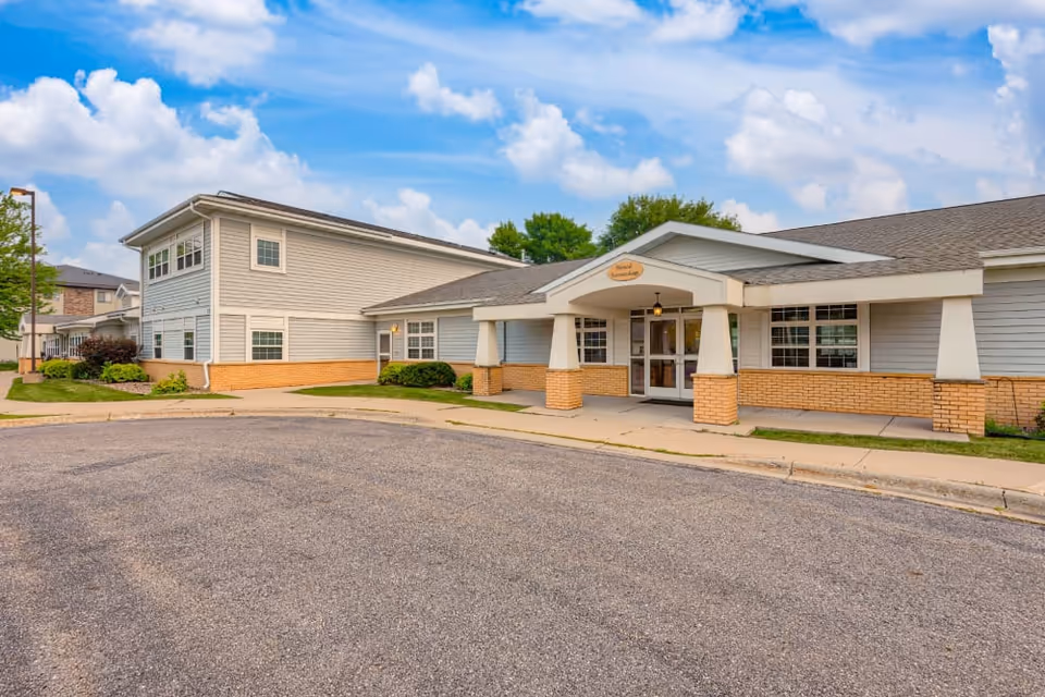 Exterior view of a single-story and two-story senior living facility building with light gray siding and brick accents, a covered entrance with pillars, and a paved driveway under a partly cloudy sky.